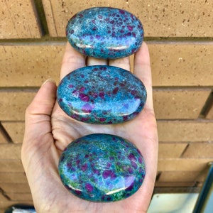 May include: Three smooth, oval-shaped stones with a blue and green base and red ruby inclusions. The stones are polished and have a glossy finish.