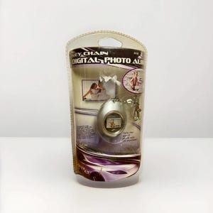 May include: A digital photo album keychain in a clear plastic package. The keychain is silver and has a small screen displaying a photo. The packaging includes the text "KEY CHAIN DIGITAL PHOTO ALBUM".