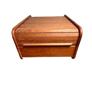 May include: A wooden bread box with a roll-top lid and a drawer. The box is made of a warm-toned wood, with a curved design on the sides and top. The box is rectangular and sits on a flat base.