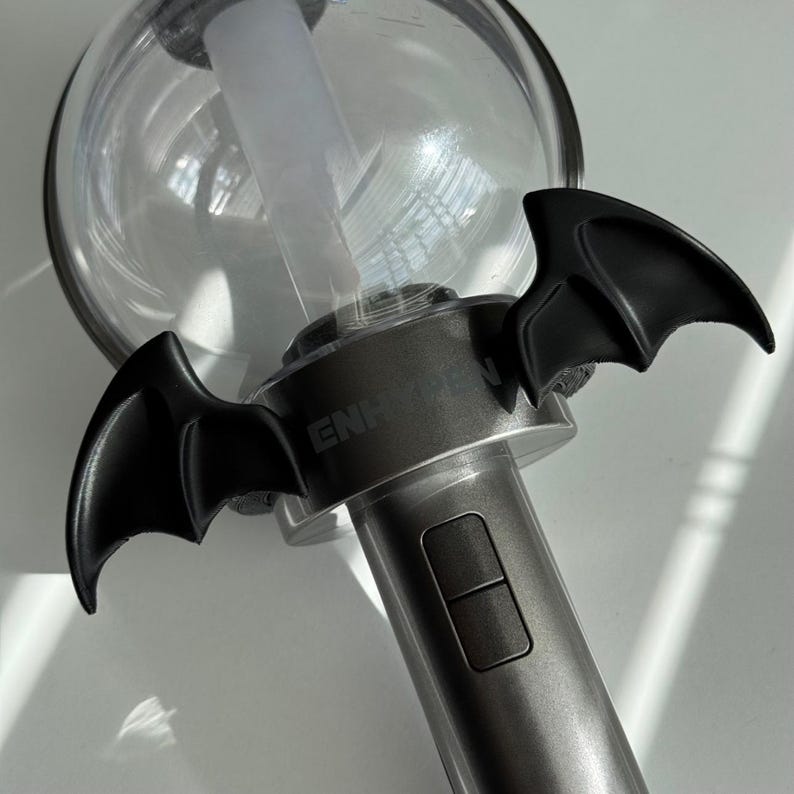 Puede incluir: Un bast&oacute;n de luz plateado con alas de murci&eacute;lago negras adjuntas. El bast&oacute;n de luz tiene una parte superior redonda y transparente y un bot&oacute;n rectangular en el mango. La palabra "ENHYPEN" est&aacute; impresa en el mango.