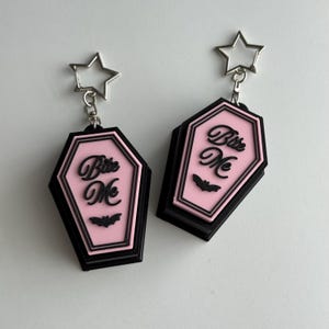 Könnte beinhalten: Ein Paar Sarg-Ohrringe mit rosa Mitte und schwarzem Rand. Die Ohrringe zeigen den Text "Bite Me" und eine Fledermaus-Grafik. Jeder Ohrring ist an einem silbernen, sternförmigen Verschluss befestigt.
