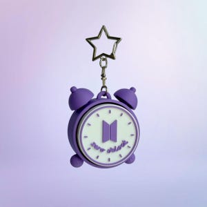 May include: A purple alarm clock keychain featuring a white clock face with the BTS logo in the center and the text zero o'clock written below it. The keychain includes a silver-toned metal star-shaped clasp attached to the top of the clock.