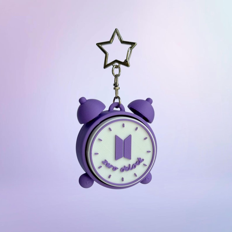 May include: A purple alarm clock keychain featuring a white clock face with the BTS logo in the center and the text zero o'clock written below it. The keychain includes a silver-toned metal star-shaped clasp attached to the top of the clock.