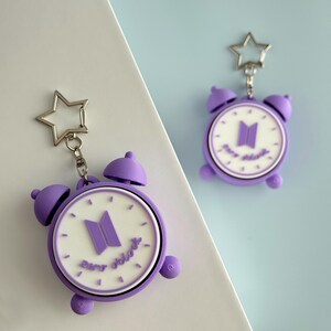 May include: Two purple and white alarm clock keychains featuring a star-shaped metal ring. The clock face is white with purple hour markers, a purple logo in the center, and the text "zero o'clock" written in purple script at the bottom.