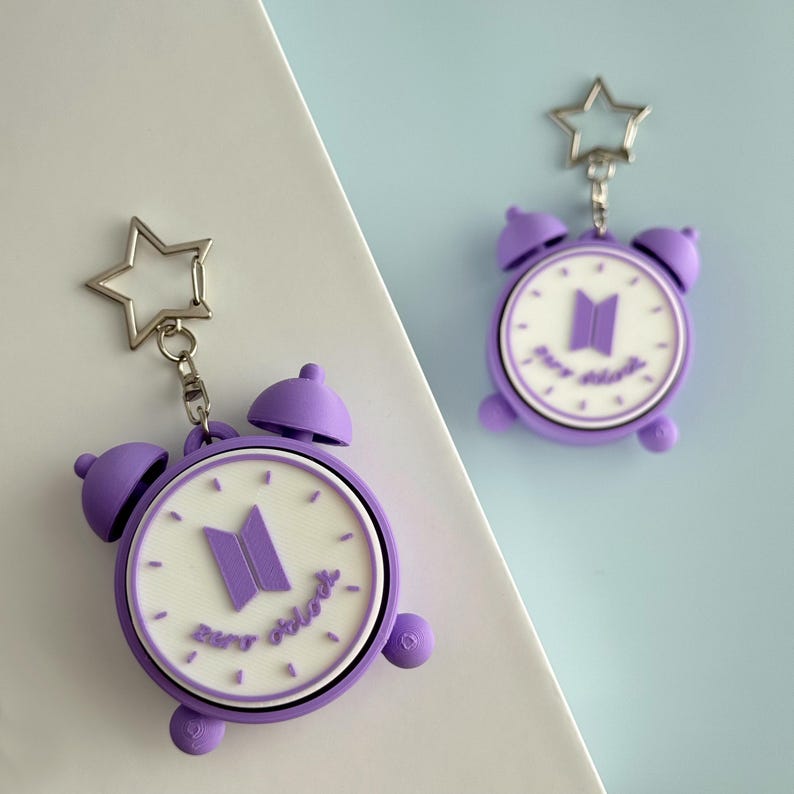 May include: Two purple and white alarm clock keychains featuring a star-shaped metal ring. The clock face is white with purple hour markers, a purple logo in the center, and the text "zero o'clock" written in purple script at the bottom.