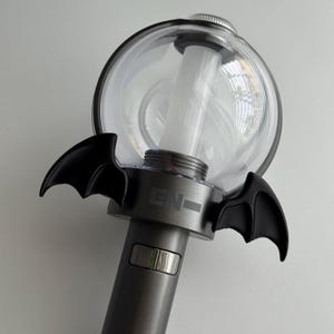 May include: A gray and clear concert light stick with black bat wings attached. The light stick has a clear, spherical top and a cylindrical handle. The letters "EN-" are printed on the side of the handle.