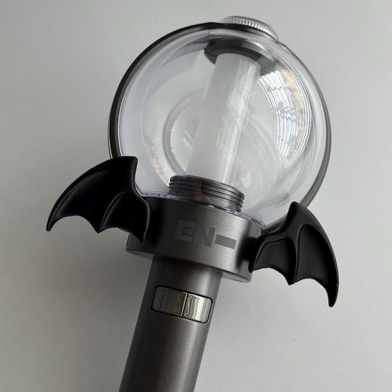 Puede incluir: Un palo de luz de concierto gris y transparente con alas de murci&eacute;lago negras. El palo de luz tiene una parte superior esf&eacute;rica transparente y un mango cil&iacute;ndrico. Las letras "EN-" est&aacute;n impresas en el lateral del mango.