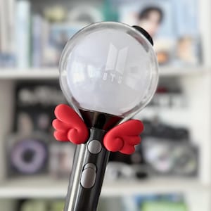 Op de afbeelding: Een zwarte en zilveren lichtstaaf met een heldere, bolvormige kop. De kop heeft het BTS-logo in het wit. Rode vleugelvormige accenten zijn bevestigd aan de basis van de kop. De lichtstaaf heeft een knop en een schakelaar op de handgreep.