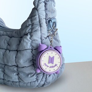 May include: A light blue quilted shoulder bag featuring a purple alarm clock keychain. The keychain has a white clock face with a purple BTS logo and the text zero o'clock printed in purple, attached by a silver star-shaped metal clasp.