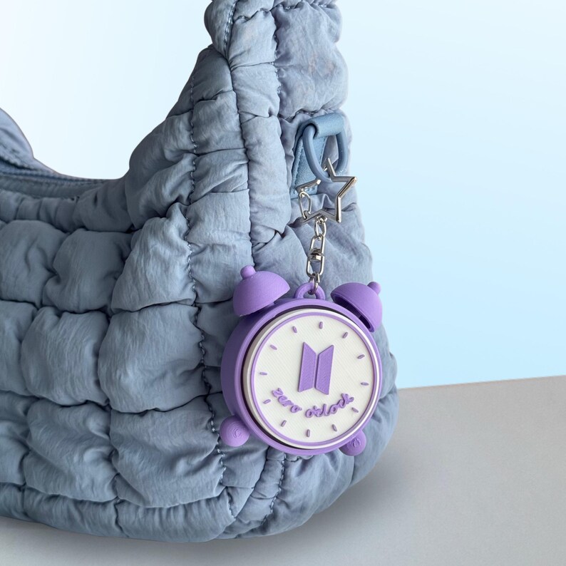 May include: A light blue quilted shoulder bag featuring a purple alarm clock keychain. The keychain has a white clock face with a purple BTS logo and the text zero o'clock printed in purple, attached by a silver star-shaped metal clasp.
