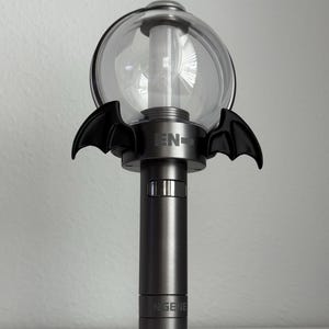 Puede incluir: Un bast&oacute;n de luz gris oscuro con una parte superior esf&eacute;rica transparente y detalles de alas de murci&eacute;lago negras. La base tiene el texto "ENGENE" impreso. El bast&oacute;n de luz est&aacute; dise&ntilde;ado para ser sostenido.