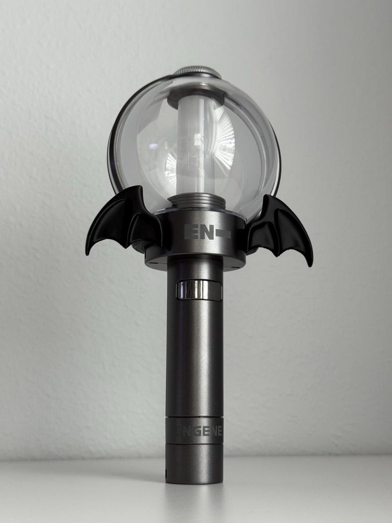 Puede incluir: Un bast&oacute;n de luz gris oscuro con una parte superior esf&eacute;rica transparente y detalles de alas de murci&eacute;lago negras. La base tiene el texto "ENGENE" impreso. El bast&oacute;n de luz est&aacute; dise&ntilde;ado para ser sostenido.