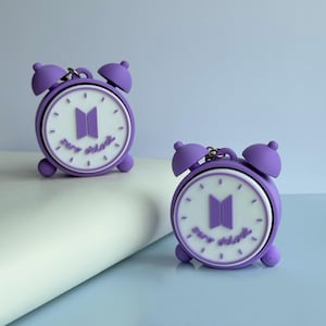 May include: Two purple silicone cases for wireless earbuds, shaped like miniature twin-bell alarm clocks. The white clock face features a purple logo consisting of two vertical rectangles and the text zero o'clock in purple script lettering.