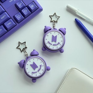 May include: Two purple alarm clock keychains with a white clock face, featuring a purple logo and the text zero o'clock. Each keychain has a silver star-shaped clasp attached to a metal chain, designed as a decorative accessory for keys or bags.