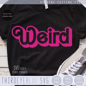 Peut inclure: Un t-shirt noir avec un graphisme rose "Weird" dans un style de police rétro. Le t-shirt est posé sur une surface en bois blanc avec une paire de chaussures en toile blanche.