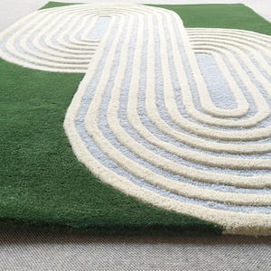 Puede incluir: Una alfombra verde con un diseño único. Presenta un gran patrón superpuesto en crema y azul claro, creando un aspecto moderno y artístico. La textura de la alfombra parece suave y afelpada, adecuada para una sala de estar o dormitorio.