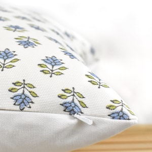 Puede incluir: Primer plano de un coj&iacute;n blanco con un estampado floral. El patr&oacute;n presenta flores azules con hojas verdes. Una cremallera blanca es visible en el lateral. El coj&iacute;n est&aacute; sobre una superficie de madera.