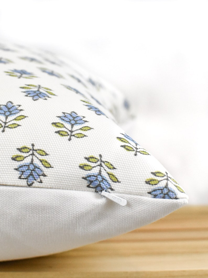 Puede incluir: Primer plano de un coj&iacute;n blanco con un estampado floral. El patr&oacute;n presenta flores azules con hojas verdes. Una cremallera blanca es visible en el lateral. El coj&iacute;n est&aacute; sobre una superficie de madera.