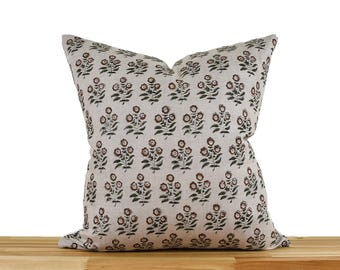 May include: A square decorative pillow with a floral pattern. The pillow features a repeating design of brown flowers with green leaves on a light beige background. The pillow is placed on a wooden surface.