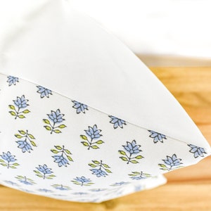 Puede incluir: Un coj&iacute;n blanco con un dise&ntilde;o floral. El patr&oacute;n presenta flores azules y hojas verdes sobre un fondo blanco. El coj&iacute;n tiene una parte superior blanca lisa.