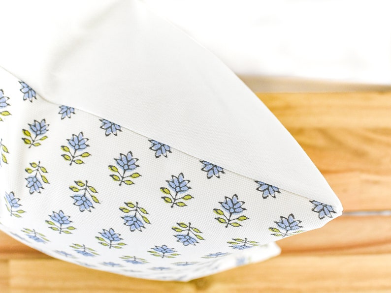 Puede incluir: Un coj&iacute;n blanco con un dise&ntilde;o floral. El patr&oacute;n presenta flores azules y hojas verdes sobre un fondo blanco. El coj&iacute;n tiene una parte superior blanca lisa.