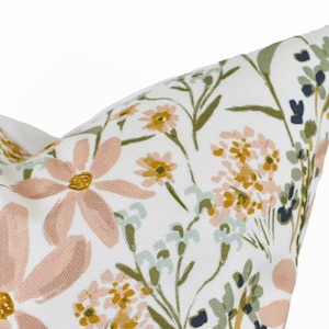 May include: Close-up of a white pillow with a floral pattern. The design features large, light pink daisies, small yellow flowers, and green and blue foliage. The pillow has a soft, textured appearance.