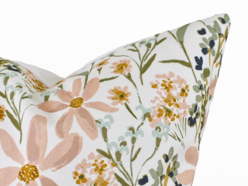 May include: Close-up of a white pillow with a floral pattern. The design features large, light pink daisies, small yellow flowers, and green and blue foliage. The pillow has a soft, textured appearance.