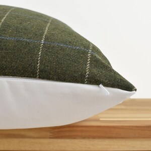 May include: Close-up of a decorative pillow with a green plaid pattern. The pillow has a white bottom and a zipper closure. The plaid design features thin white and blue lines on a green background.