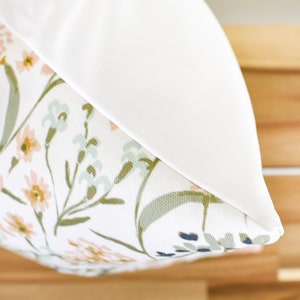 May include: A decorative pillow with a floral pattern. The pillow features a white background with a design of pink, yellow, and blue flowers with green stems and leaves. The pillow has a solid white border.