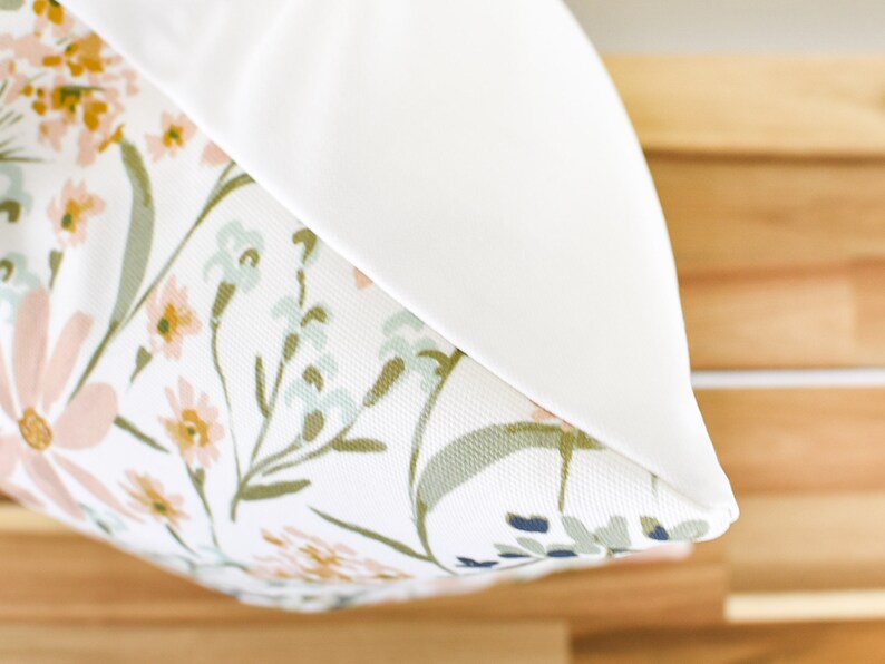 May include: A decorative pillow with a floral pattern. The pillow features a white background with a design of pink, yellow, and blue flowers with green stems and leaves. The pillow has a solid white border.