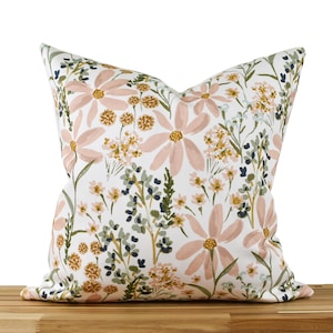 May include: Square throw pillow with a floral pattern. The pillow features large, light pink flowers with yellow centers, surrounded by green and yellow foliage on a white background. The pillow is resting on a wooden surface.