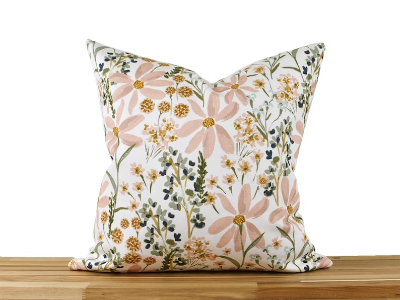 May include: Square throw pillow with a floral pattern. The pillow features large, light pink flowers with yellow centers, surrounded by green and yellow foliage on a white background. The pillow is resting on a wooden surface.