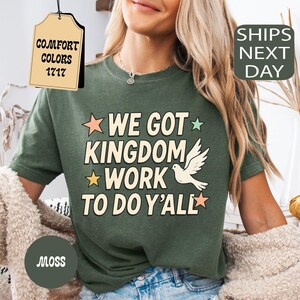 May include: Moss green t-shirt with the text "WE GOT KINGDOM WORK TO DO Y'ALL" in white, yellow, and pink. A white dove graphic is featured in the center. The shirt is a Comfort Colors 1717 style.