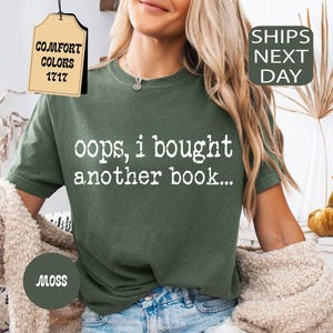 May include: A moss green t-shirt with the text "oops, i bought another book..." in white. The shirt is part of the Comfort Colors 1717 collection. The model is wearing denim shorts and a beige cardigan.