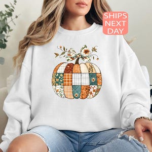 May include: White sweatshirt featuring a patchwork pumpkin design with floral and plaid patterns in orange, green, and brown. The pumpkin has a floral bow on top. The text "SHIPS NEXT DAY" is in a pink box.