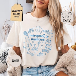 K&ouml;nnte beinhalten: Elfenbeinfarbenes T-Shirt mit einer blauen Grafik von Fitnessger&auml;ten und dem Schriftzug "movement is medicine". Das Shirt wird mit Jeansshorts getragen. Das Bild enth&auml;lt auch eine beige Strickjacke, ein dekoratives Kissen und einen Weidenkorb.