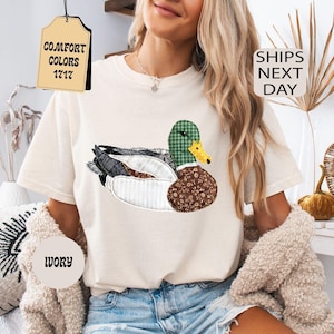 May include: Ivory-colored t-shirt featuring a patchwork duck design with green, brown, and black patterns. The shirt has short sleeves and is worn with denim shorts. The tag reads "Comfort Colors 1717".