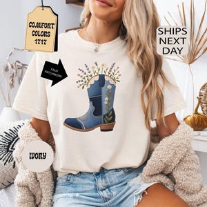 May include: An ivory short-sleeved t-shirt featuring a graphic of a blue denim cowboy boot filled with colorful wildflowers. The boot design includes faux patchwork details and floral embroidery. The shirt has a crew neckline and a relaxed fit.