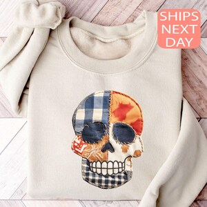Könnte beinhalten: Beiges Sweatshirt mit einem Totenkopf-Design aus Patchwork-Stoff in blauen, orangefarbenen und weißen Mustern. Der Totenkopf hat ein detailliertes, zahnreiches Grinsen. Der Text "SHIPS NEXT DAY" befindet sich in einer rosa Box.