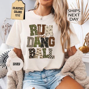 Op de afbeelding: Een ivoorkleurig t-shirt met korte mouwen en de tekst RUN THE DANG BALL in een blokkerig lettertype met meerdere patronen. De letters bevatten diverse ontwerpen met een football-thema, waaronder strepen, ruitjes en kleine football-iconen.