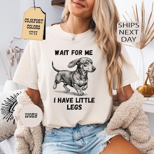 May include: Ivory-colored t-shirt featuring a dachshund illustration with the text "WAIT FOR ME I HAVE LITTLE LEGS." The shirt has a classic crew neck and short sleeves. The image also includes tags that say "Comfort Colors 1717" and "Ivory."