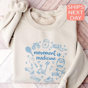 May include: Beige sweatshirt with the phrase "movement is medicine" in blue, surrounded by blue illustrations of fitness equipment. A scrunchie is on the shoulder. The sweatshirt is folded on a wooden surface.