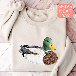 May include: Beige sweatshirt with an appliqued duck design. The duck has a green plaid head, a brown floral body, and a yellow beak. The sweatshirt is folded, with the text "SHIPS NEXT DAY" in a pink box.