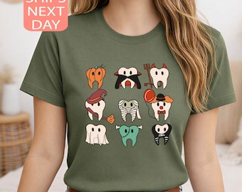 May include: Olive green t-shirt featuring a Halloween-themed graphic of cartoon teeth dressed in costumes. The design includes a pumpkin, vampire, ghost, and other spooky characters. The shirt also has the text "SHIPS NEXT DAY".