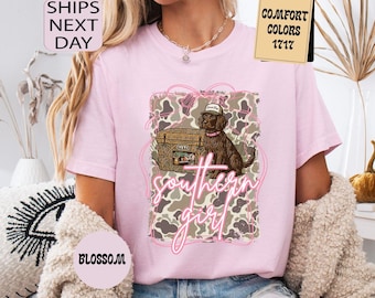 May include: A light pink t-shirt featuring a graphic of a brown dog wearing a cap, set against a camouflage background. The words "southern girl" are written in a pink script font. The shirt has the text "Comfort Colors 1717".