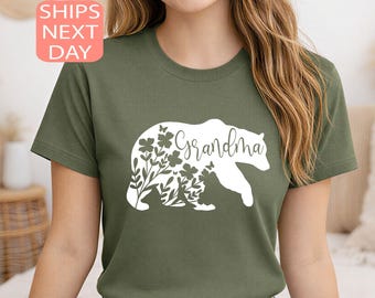 Puede incluir: Camiseta verde oliva con una silueta de oso blanco llena de diseños florales y mariposas. La palabra "Grandma" está escrita en cursiva sobre el oso. La camiseta tiene cuello redondo y mangas cortas.