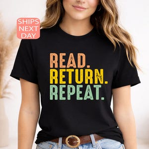 以下が含まれることがあります： オレンジ、黄色、緑で「READ. RETURN. REPEAT.」と書かれた黒いTシャツ。 シャツはクルーネックと半袖です。 モデルはブルージーンズと茶色のベルトを着用しています。