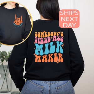 May include: Black sweatshirt with a retro design on the back that reads "SOMEBODY'S TIRED ASS MILK MAKER" in orange, pink, and blue. The front features a skeleton hand graphic with text. A fun, casual top.