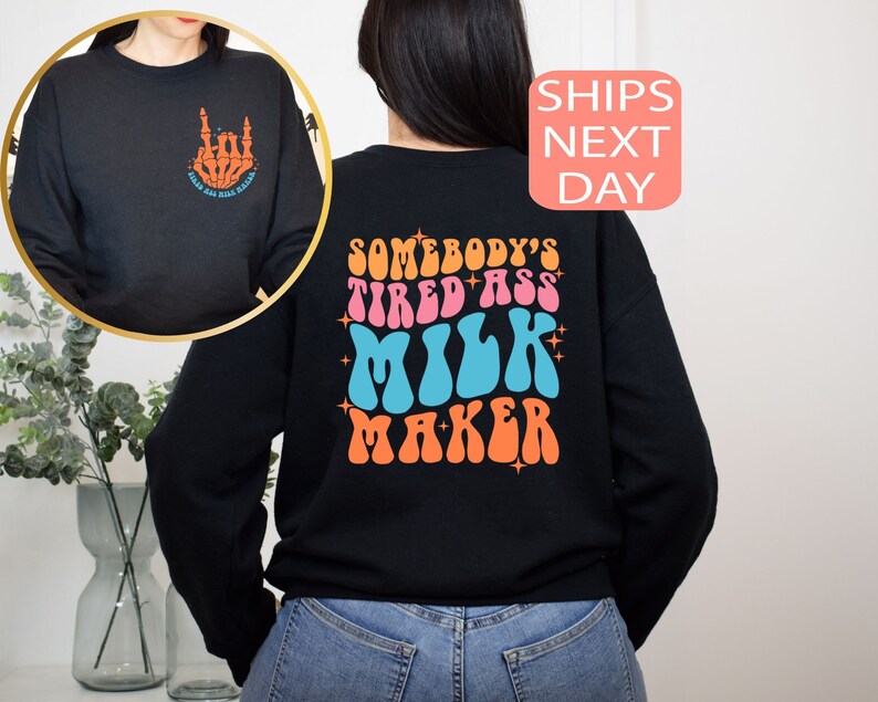 May include: Black sweatshirt with a retro design on the back that reads "SOMEBODY'S TIRED ASS MILK MAKER" in orange, pink, and blue. The front features a skeleton hand graphic with text. A fun, casual top.