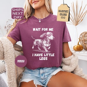 May include: Berry-colored t-shirt with a dachshund graphic and the text "WAIT FOR ME I HAVE LITTLE LEGS." The shirt is a Comfort Colors 1717 style. The model is wearing denim shorts and a cream-colored cardigan.